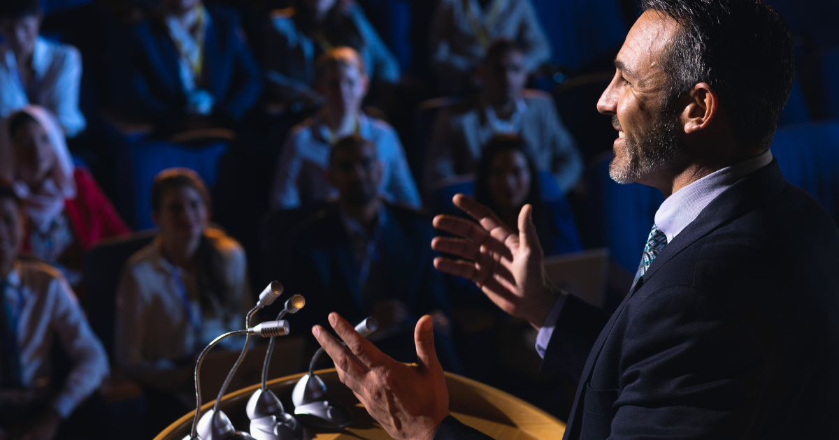 A politician stands at a podium with his hands raised while giving a speech to a crowd.
