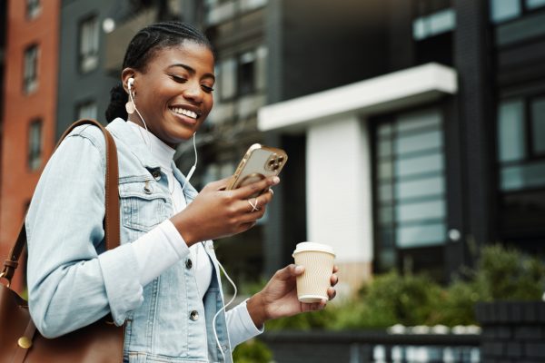 A young woman walks down the street listening to programmatic audio on a podcast while enjoying coffee.